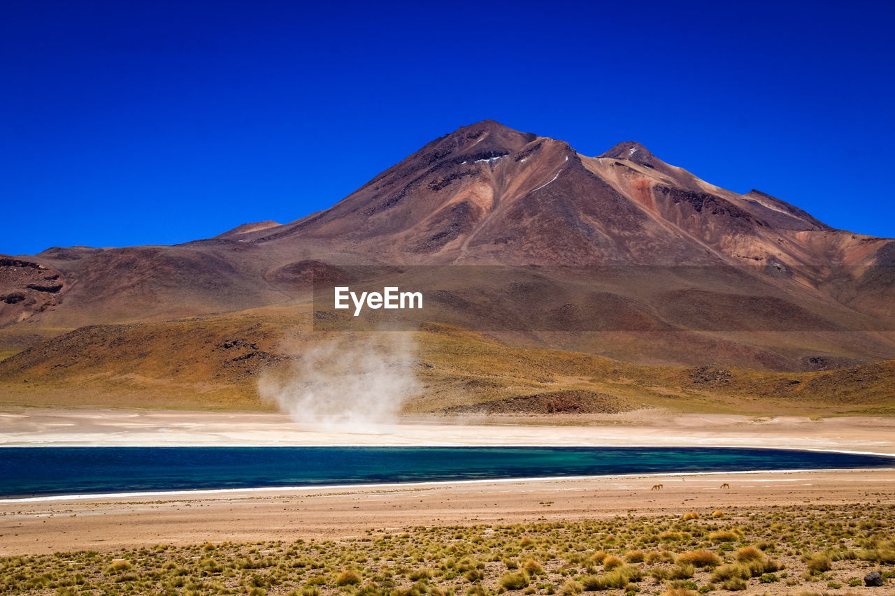 Scenic view of arid landscape with salt lake and sand storm against clear blue sky - atacama desert