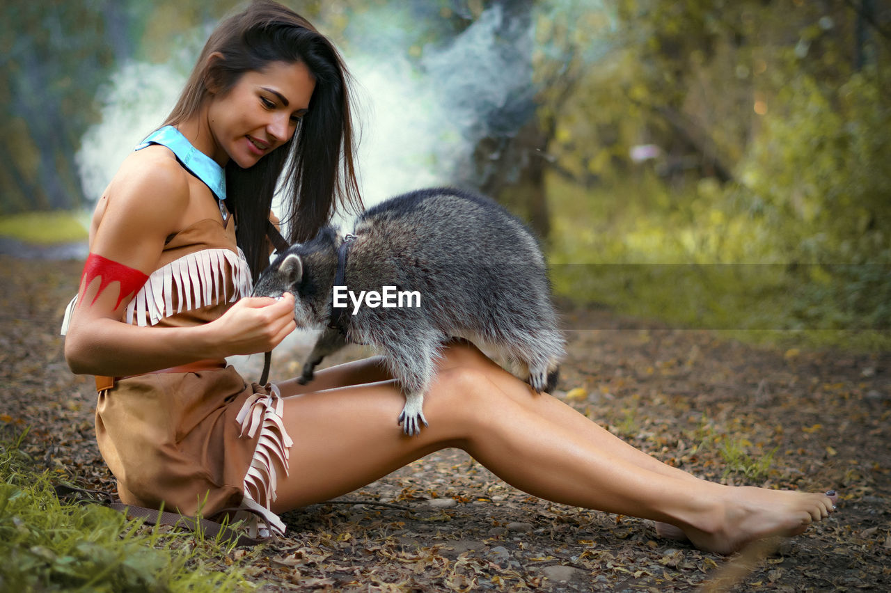 Young woman in traditional clothing playing with raccoon in forest