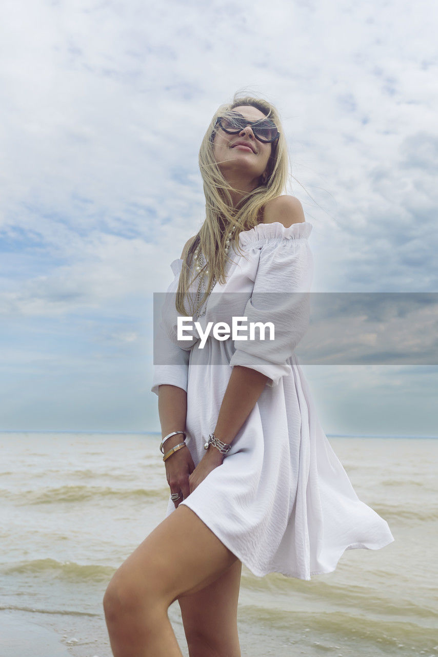 one person, women, sea, adult, water, blond hair, female, young adult, beach, land, photo shoot, fashion, clothing, sky, dress, long hair, white, hairstyle, nature, cloud, portrait, leisure activity, standing, three quarter length, holiday, vacation, day, trip, smiling, human leg, person, horizon over water, looking at camera, lifestyles, beauty in nature, spring, happiness, horizon, outdoors, limb, sand, front view, summer, emotion, full length, looking, carefree, casual clothing, glasses, wind