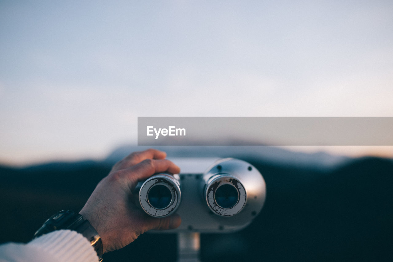 Cropped hand of man holding coin-operated binoculars against sky