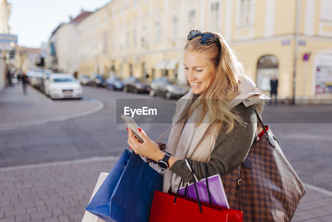 Smiling woman with shopping bags using mobile phone on street