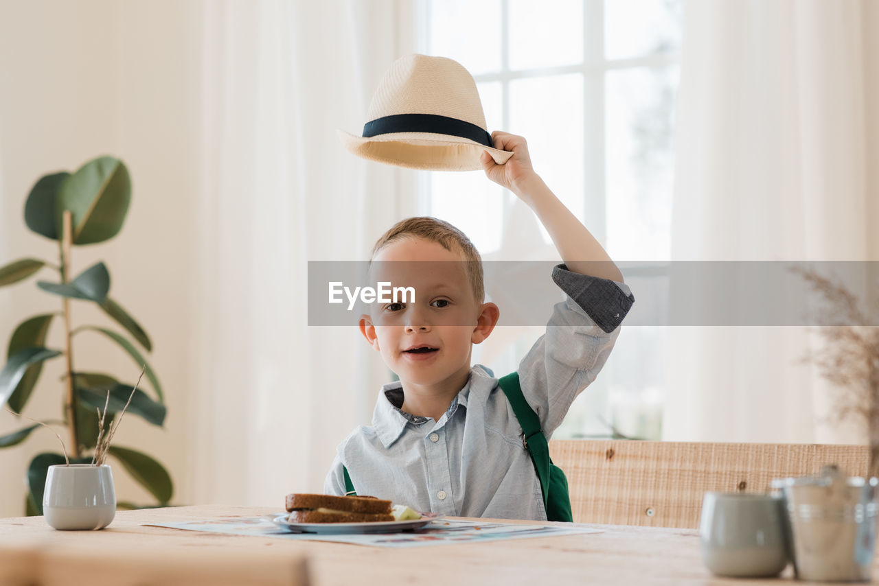 Boy holding his hat up whilst eating his lunch at home smiling