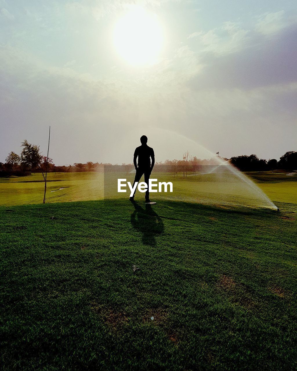 Silhouette man standing on grassy field by agricultural sprinkler during sunny day