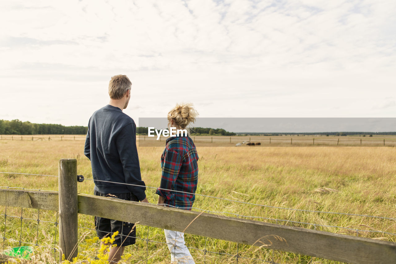 Rear view of couple standing by fence on grassy field against sky