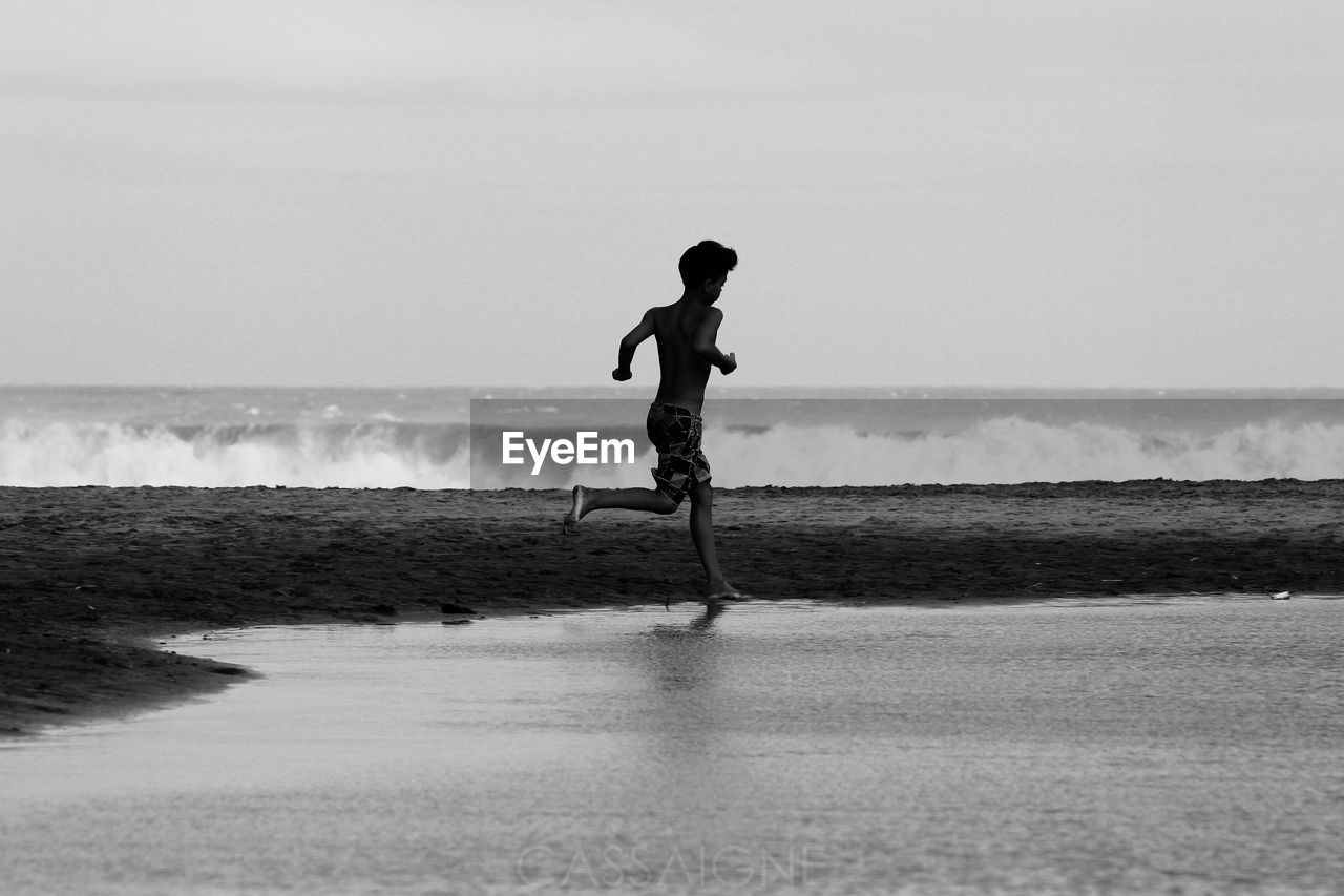 Side view of shirtless boy running at beach against sky