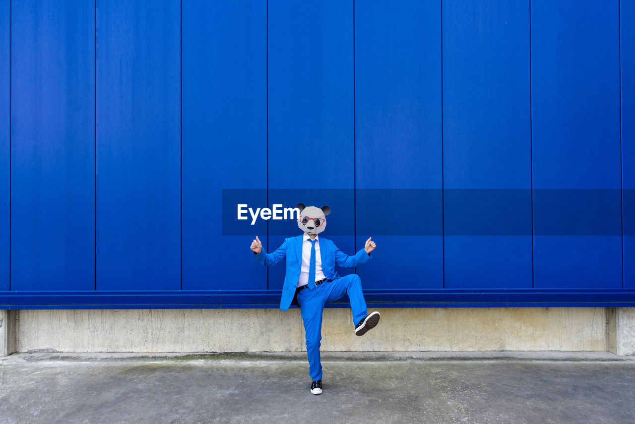 Man wearing vibrant blue suit and panda mask standing on one leg against blue wall