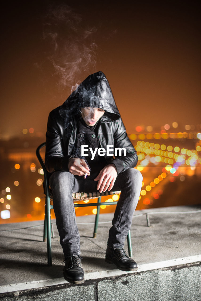 Full length of man smoking cigarette while sitting on chair at building terrace during night