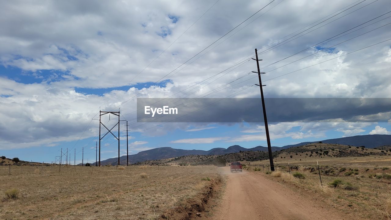 sky, electricity, cloud, landscape, environment, cable, technology, power generation, road, electricity pylon, nature, power supply, power line, land, mountain, scenics - nature, transportation, field, no people, beauty in nature, the way forward, overhead power line, dirt road, wind, rural scene, prairie, non-urban scene, desert, horizon, telephone pole, day, outdoors, dirt, remote, tranquility, blue, climate, diminishing perspective, vanishing point, tranquil scene, rural area, horizon over land, wind farm, telephone line, travel, mountain range, environmental conservation, soil