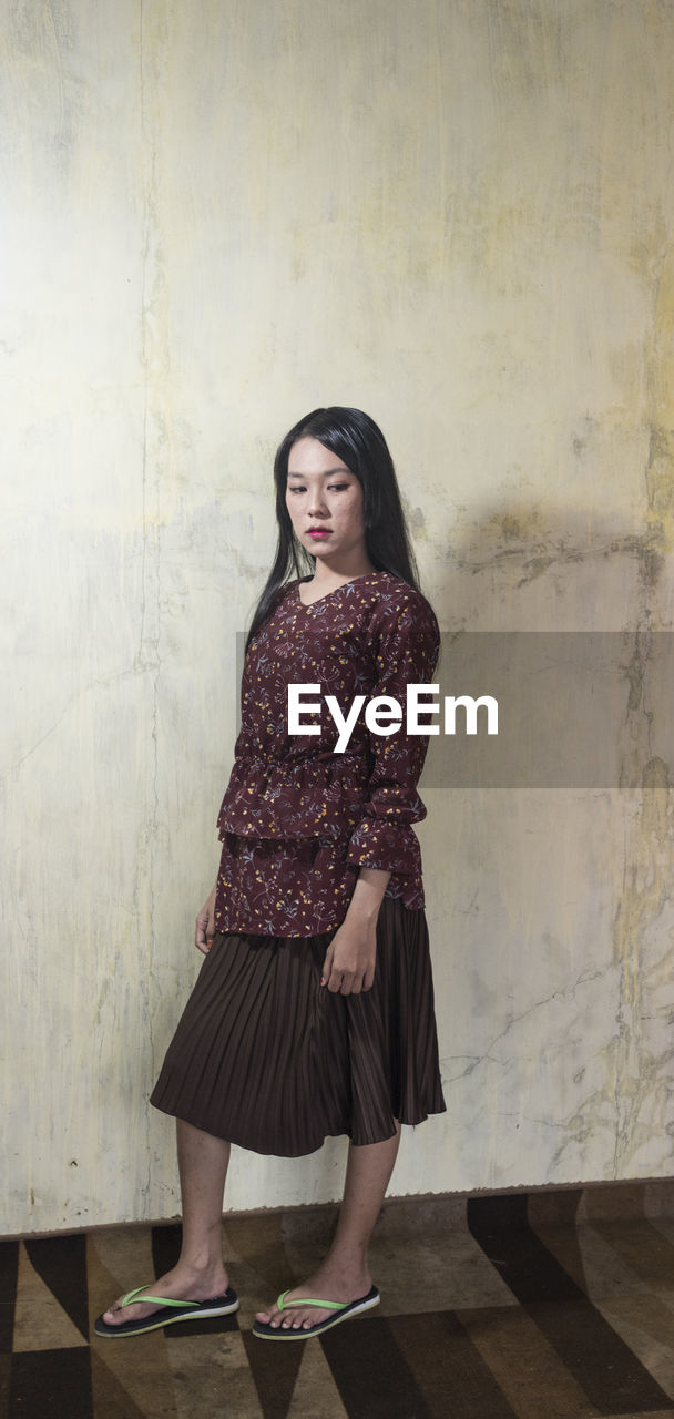 FULL LENGTH PORTRAIT OF A WOMAN STANDING AGAINST WALL