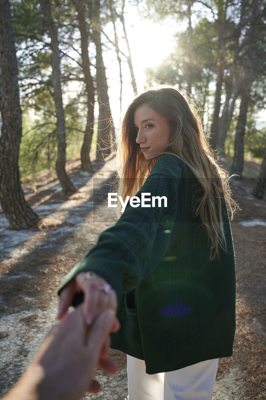 Back view of young long haired female in warm jacket holding hand of anonymous boyfriend while walking together in sunny autumn forest