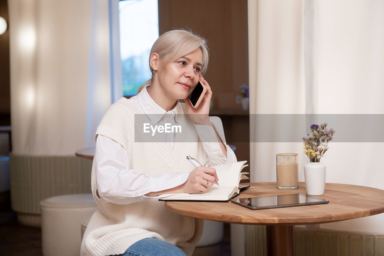 A cute girl is sitting in a cafe and talking on the phone and making notes in a diary