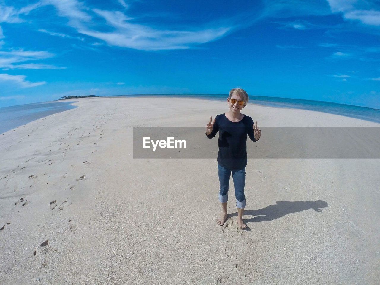 Fish-eye view of smiling man gesturing peace sign while standing on shore at beach against sky
