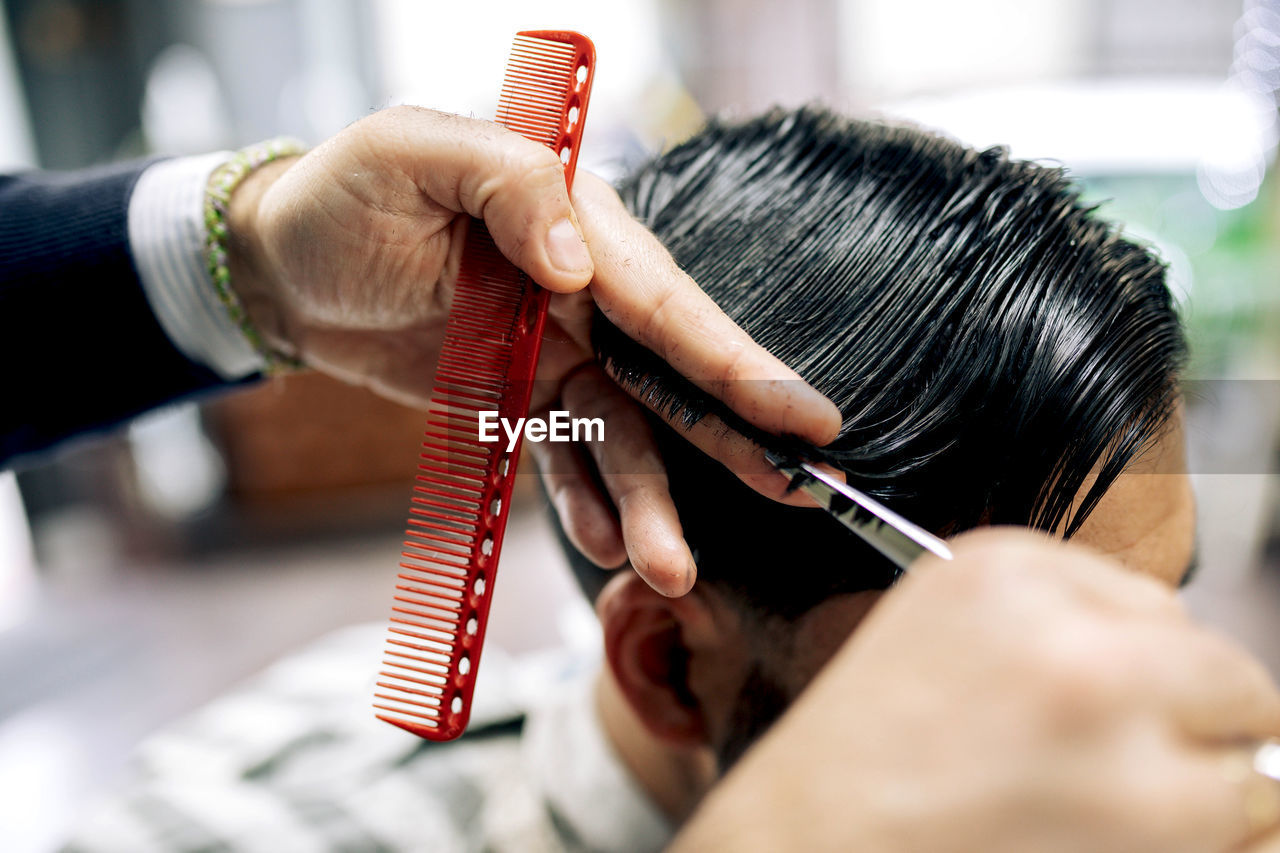 Crop anonymous barber using scissors and comb to cut hair of male client while working in grooming salon