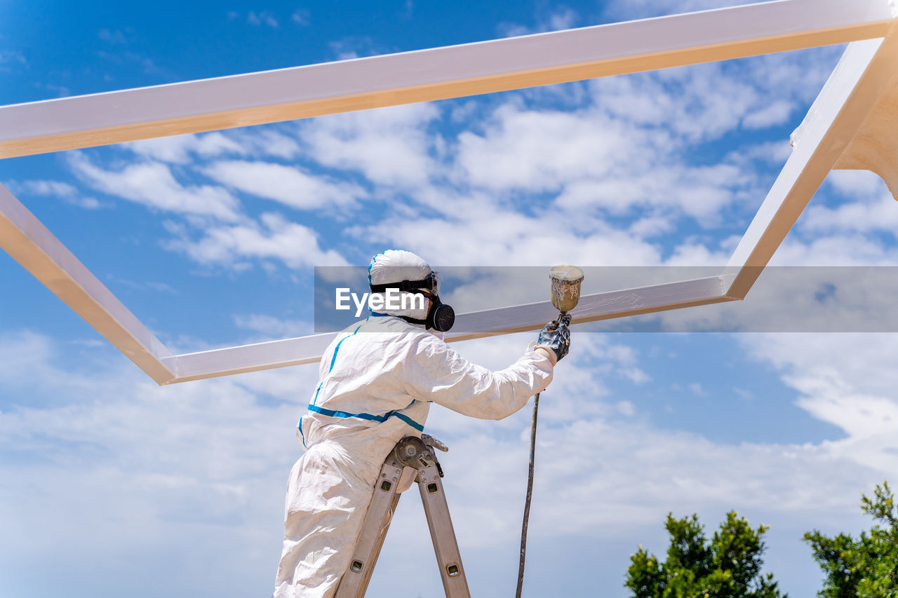 Low angle of anonymous employee in protective uniform and mask standing on ladder and using air brush to paint frame structure against cloudy sky on sunny day