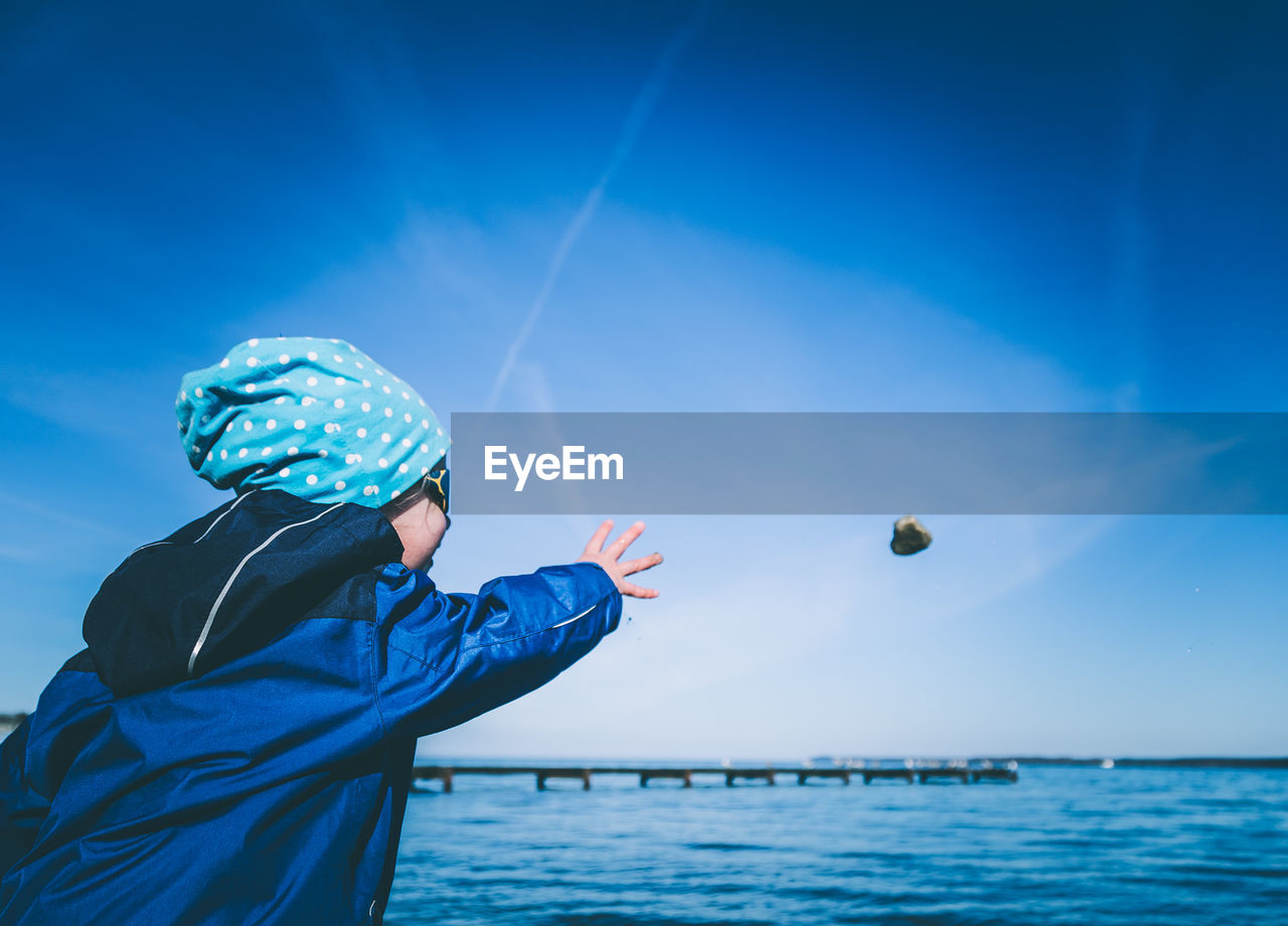 Child throwing rock in sea against blue sky