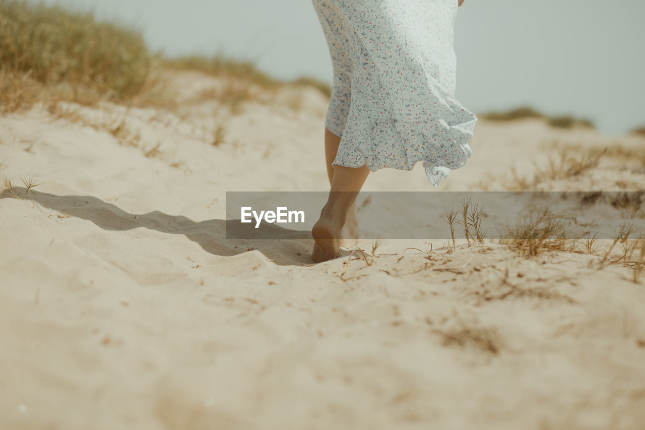 Back view of crop unrecognizable barefoot female in white summer dress walking alone on sandy beach in sunny summer day in spain