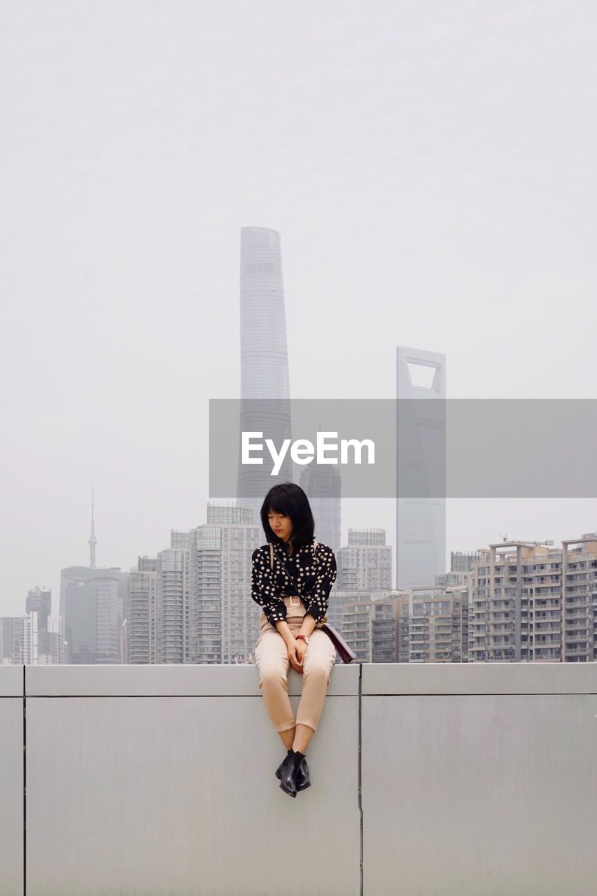 Woman sitting on wall in city against buildings and sky