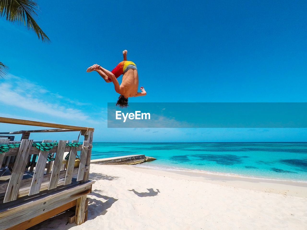 REAR VIEW OF MAN JUMPING AT BEACH AGAINST BLUE SKY