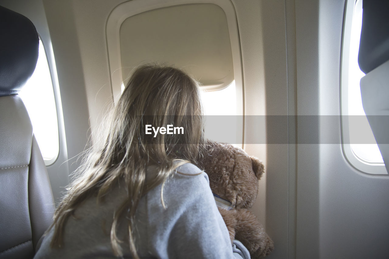 Rear view of girl with teddy bear looking through window while traveling in airplane