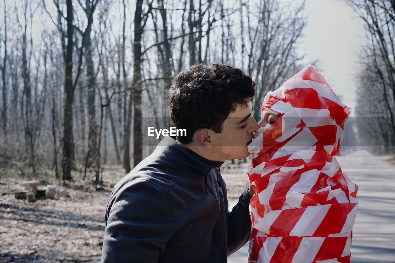Side view of man kissing woman wrapped in cordon tape standing in forest