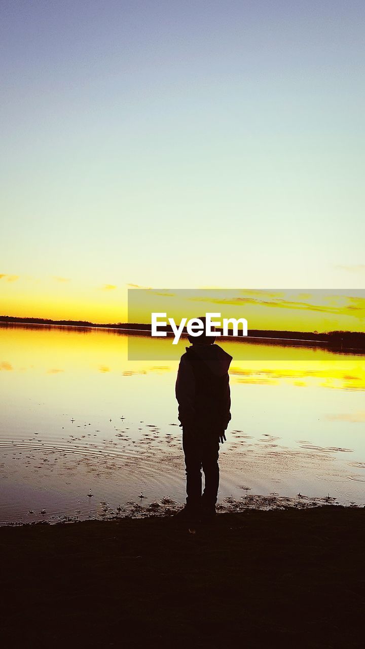 Silhouette boy standing on lakeshore against sky during sunset