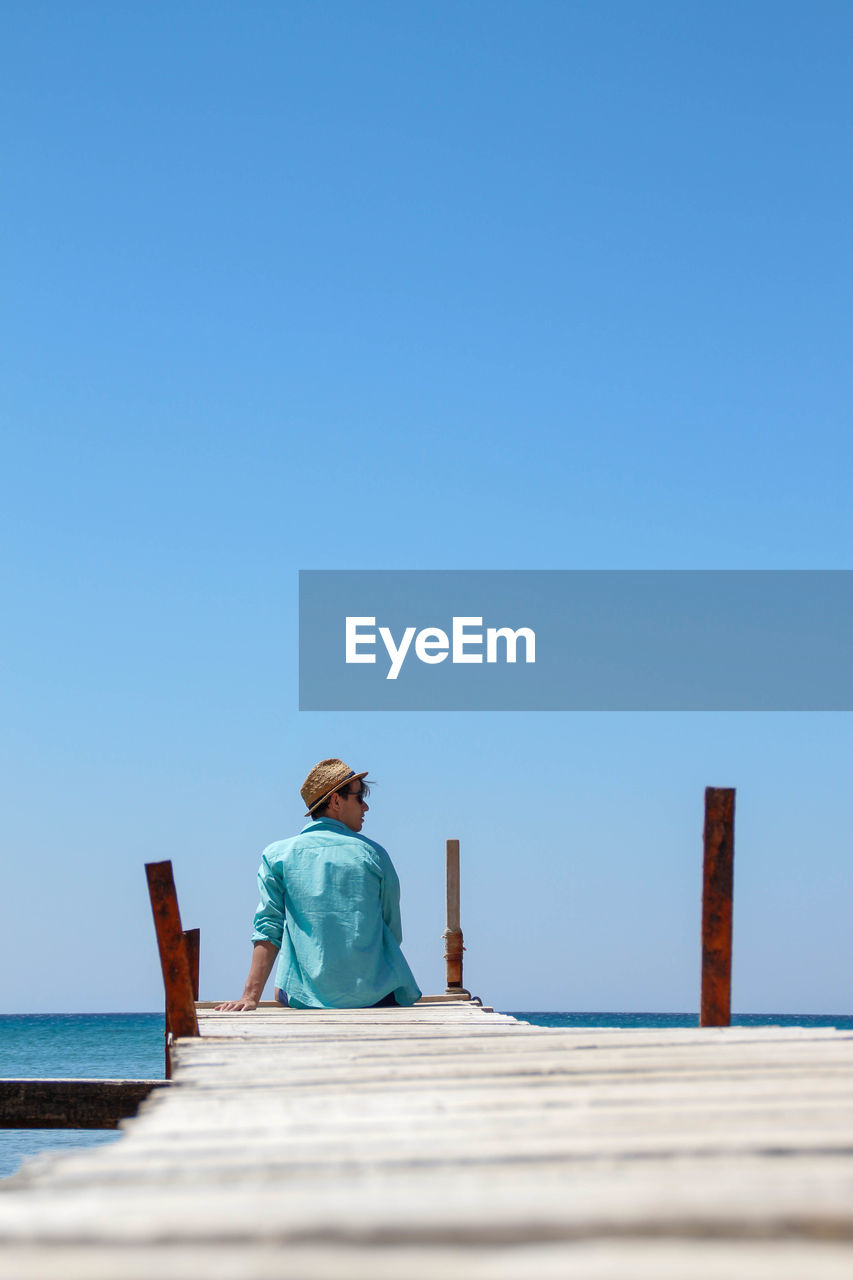 Boy dressed in a turquoise shirt sitting on an antique wooden catwalk over the mediterranean sea