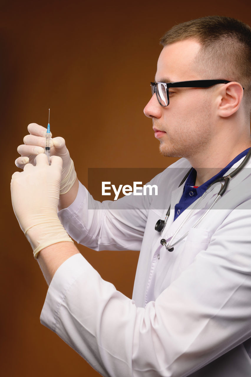 MIDSECTION OF MAN HOLDING EYEGLASSES WHILE STANDING AGAINST WALL