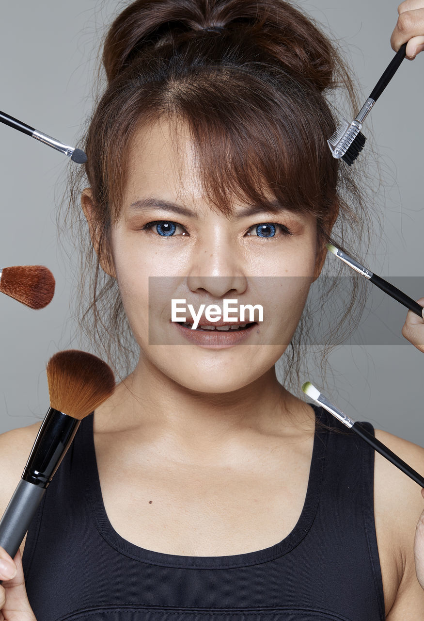 Close-up portrait of smiling beautiful woman with various make-up brushes and mascara applicator against gray background