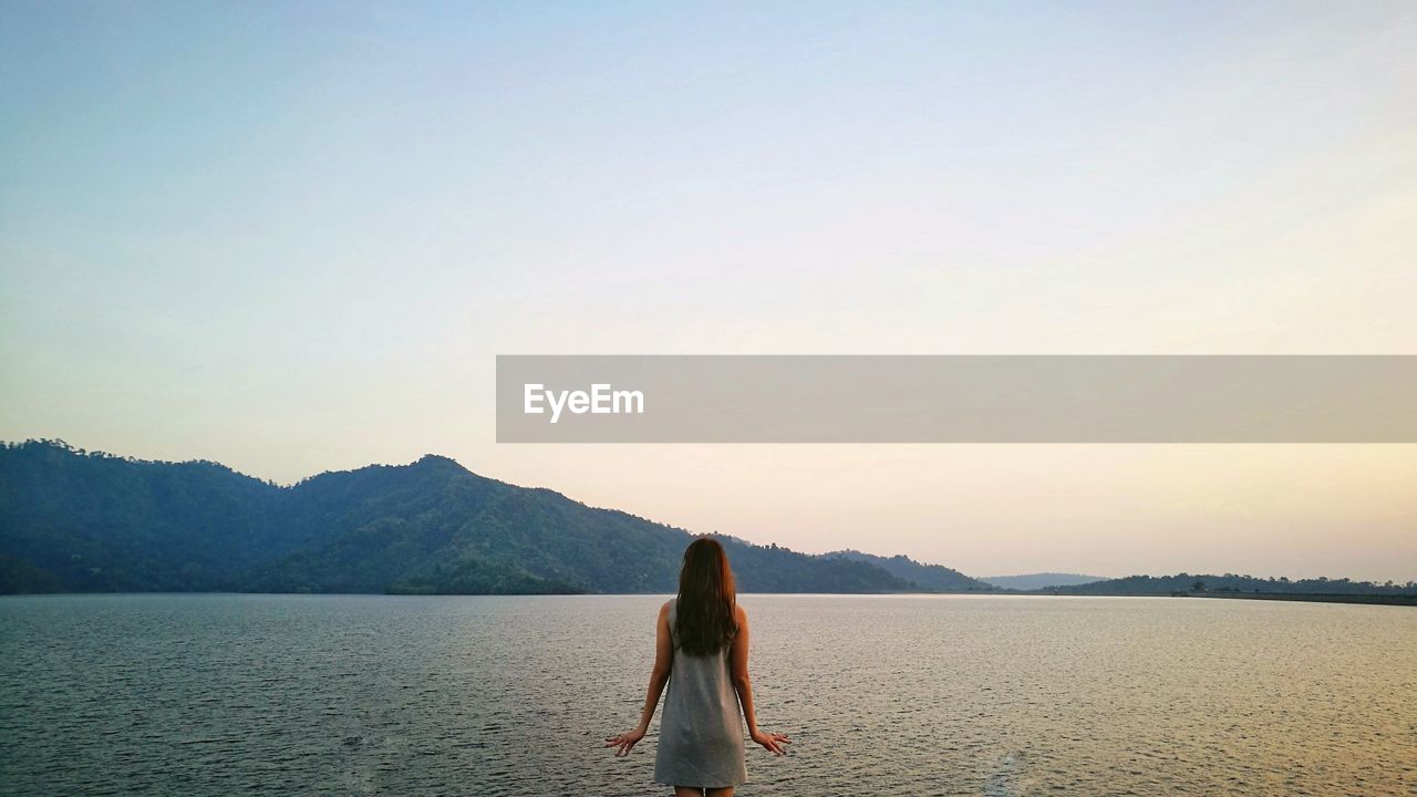 Rear view of woman looking at sea while standing against clear sky during sunset