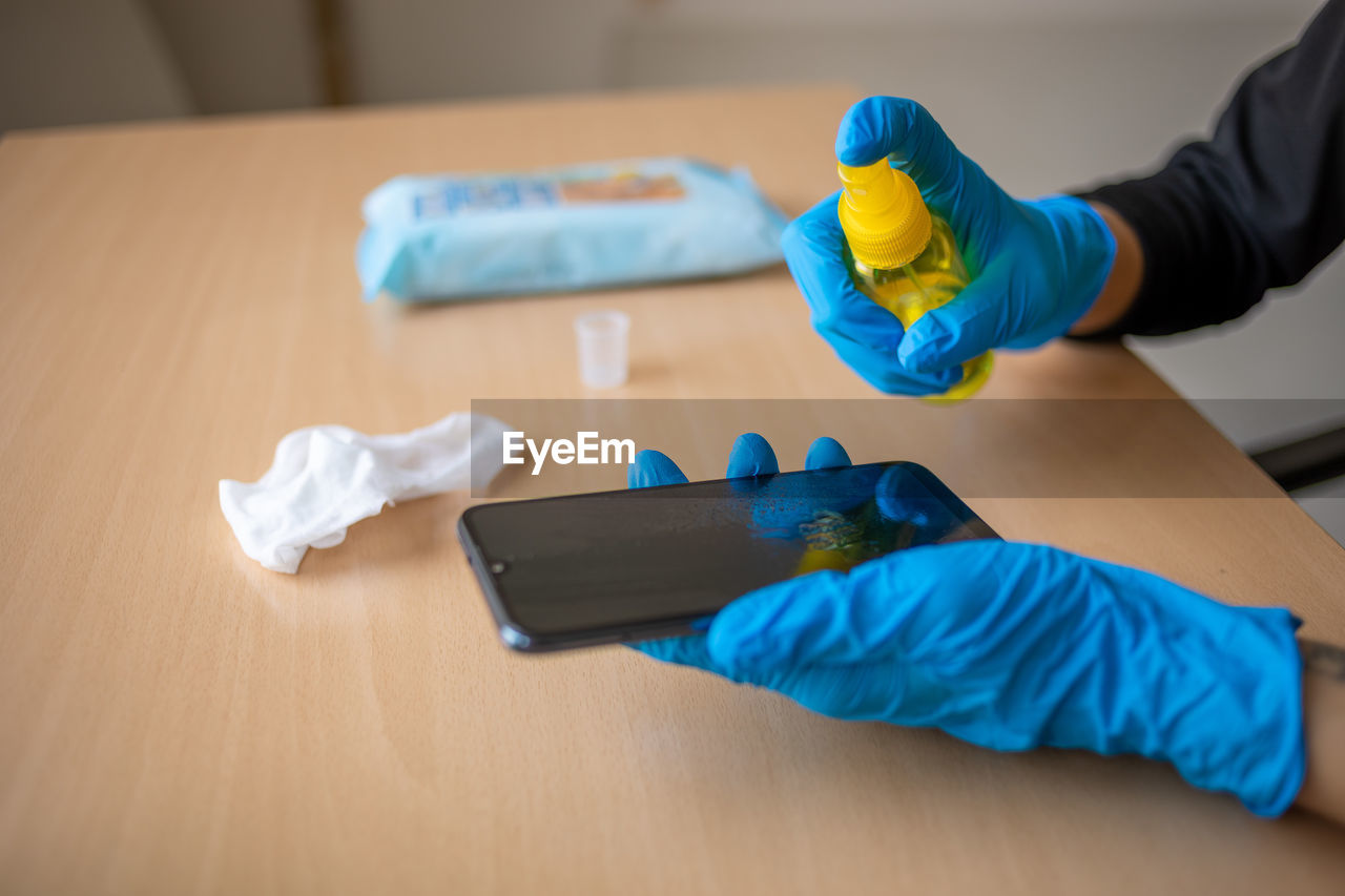 Hand in gloves sanitizing cleaning smartphone mobile phone on table surface with wet wipes alcohol