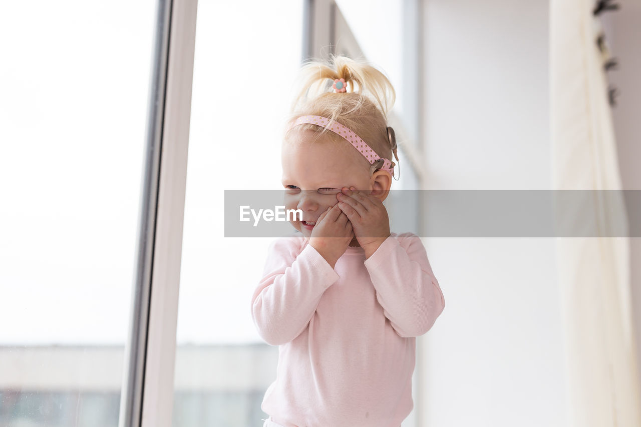 childhood, one person, child, indoors, blond hair, toddler, window, innocence, women, baby, emotion, looking, female, day, portrait, standing, waist up, cute, clothing, copy space, lifestyles, person, home interior, casual clothing, happiness, domestic life, front view, smiling, pink, human face, hairstyle, dress, adult