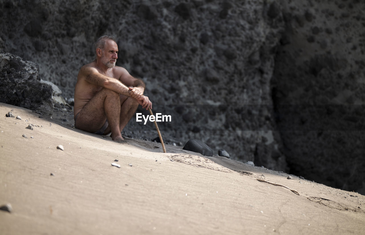 Full length of shirtless adult man in swimwear sitting on beach against rock