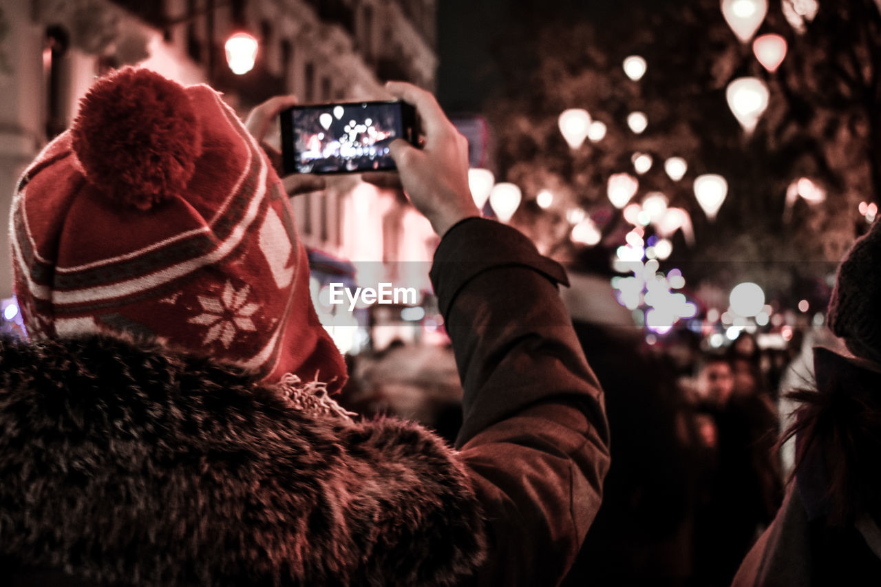 Rear view of woman photographing through mobile phone on street at night