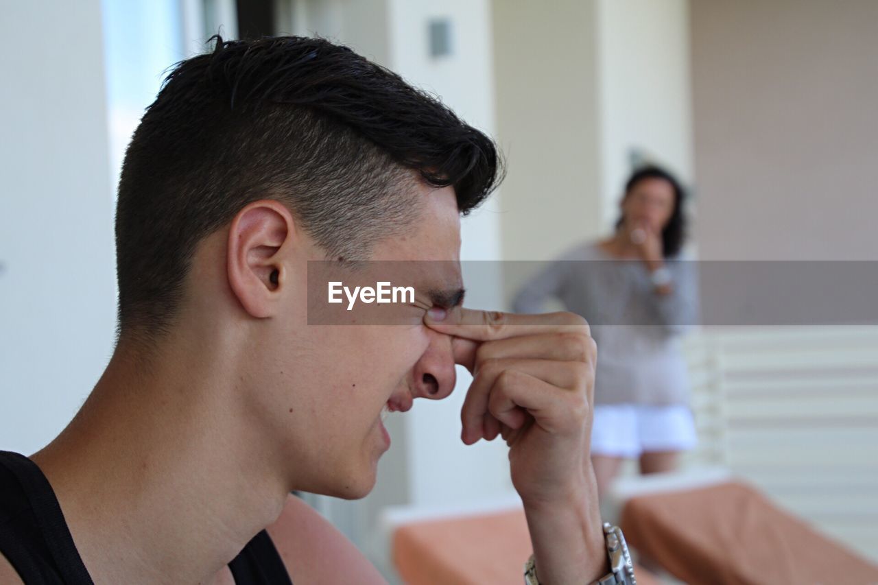 Young man rubbing eyes while mother standing by wall at home