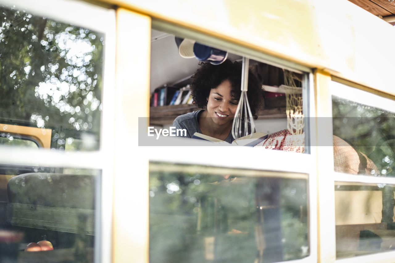 Young afro woman reading book while lying in motor home seen through window