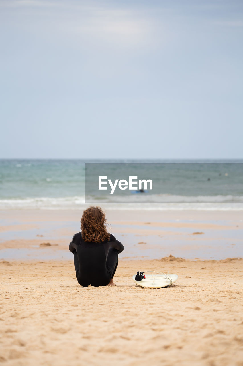 Rear view of man sitting by surfboard at beach