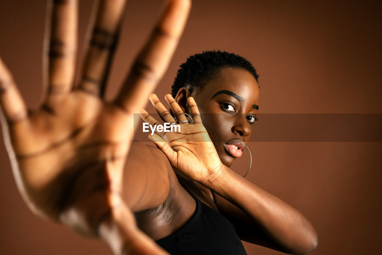 Black female model with short hair looking at camera and making protective gesture against brown background