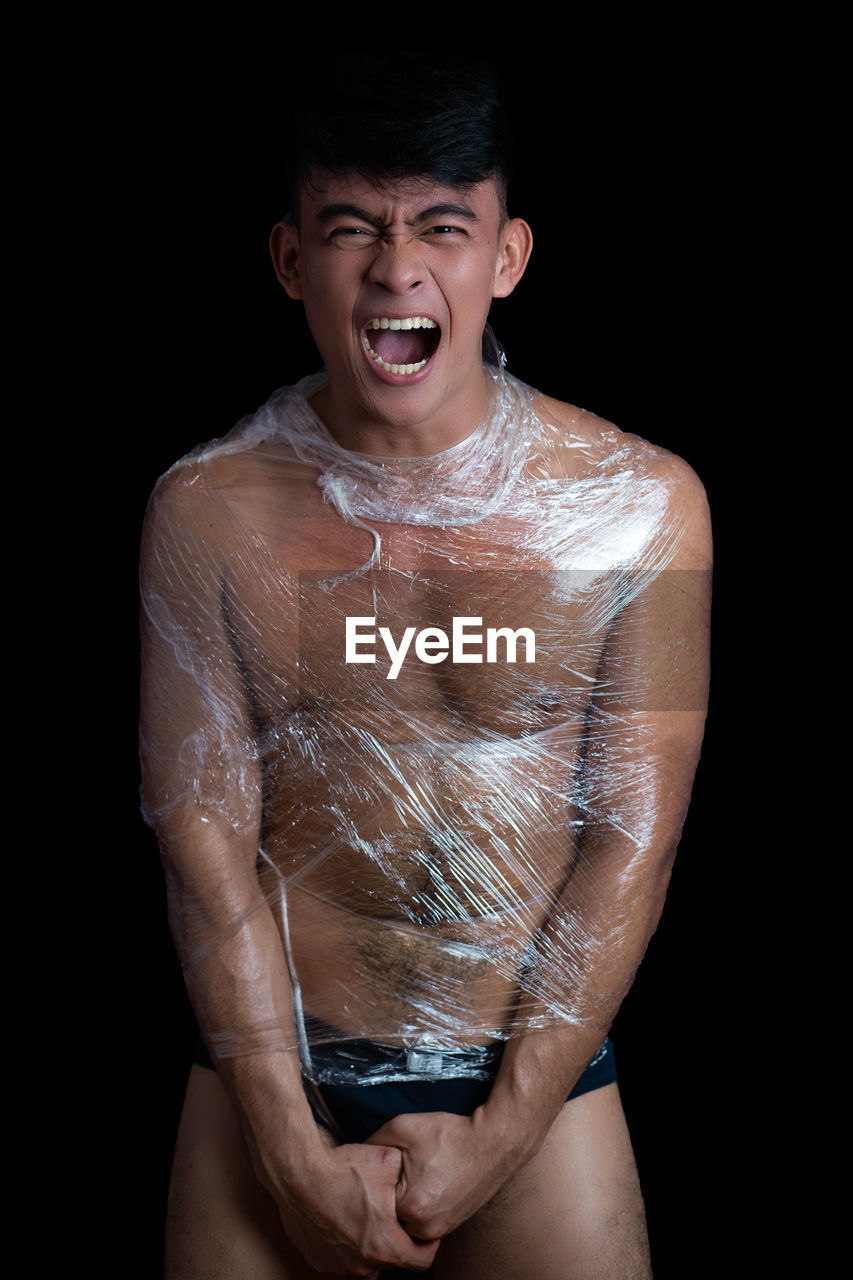 Portrait of shirtless young man wrapped in plastic against black background