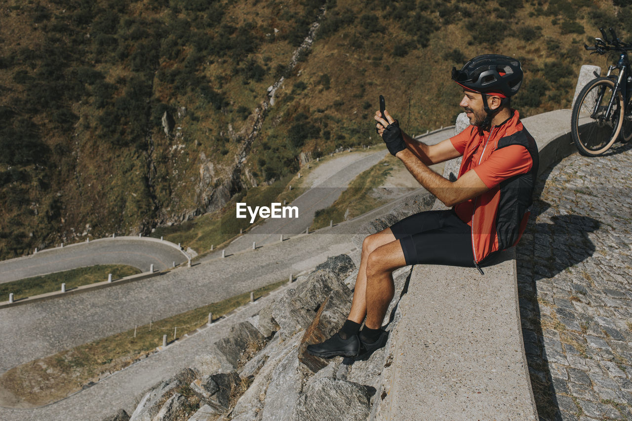 Cyclist photographing through smart phone sitting on wall at gotthard pass