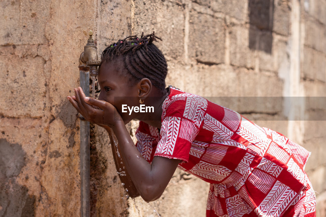 Girl drinking water from faucet against wall