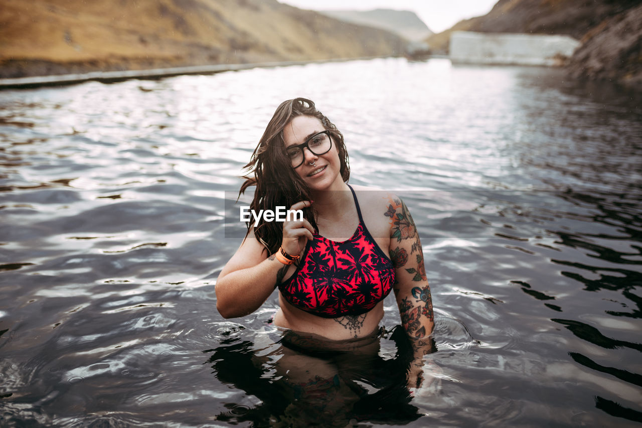 Young cheerful tattooed hipster in swimsuit in water between mountains