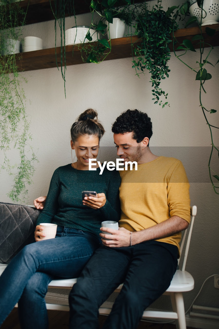 Smiling boyfriend and girlfriend looking at mobile phone while having coffee in living room