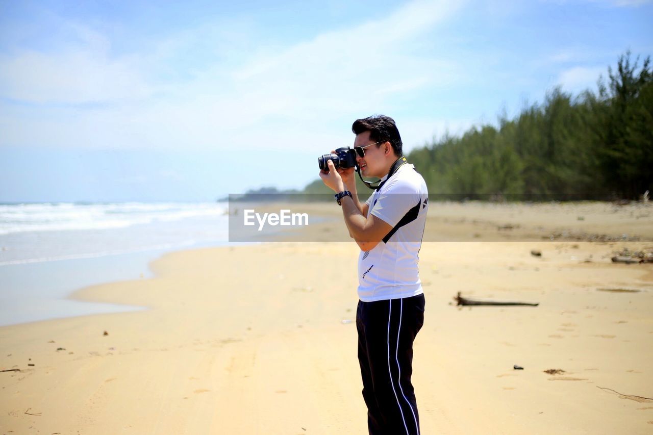 Man photographing through camera while standing on sand at beach