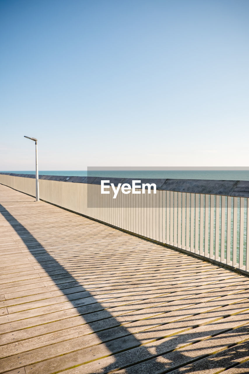 sky, walkway, boardwalk, water, sea, nature, horizon, clear sky, sunlight, tranquility, beach, horizon over water, scenics - nature, land, beauty in nature, sunny, blue, tranquil scene, no people, coast, day, pier, footpath, copy space, wood, line, ocean, shore, outdoors, architecture, railing, environment, travel destinations, the way forward, travel, summer, absence, landscape, shadow, sand, urban skyline, built structure, holiday, tourism, trip, idyllic, vacation, empty