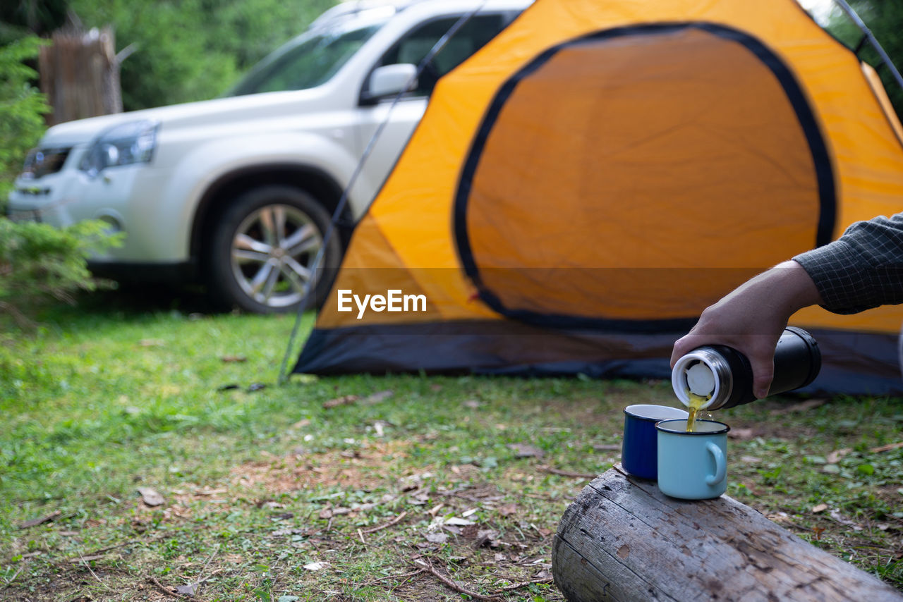 Man hand pour tea in metal cup. yellow tent with suv on background. union with nature