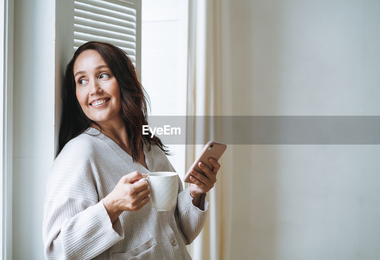 Young woman in cozy cardigan with cup of tea in hands using mobile phone in bright interior at home