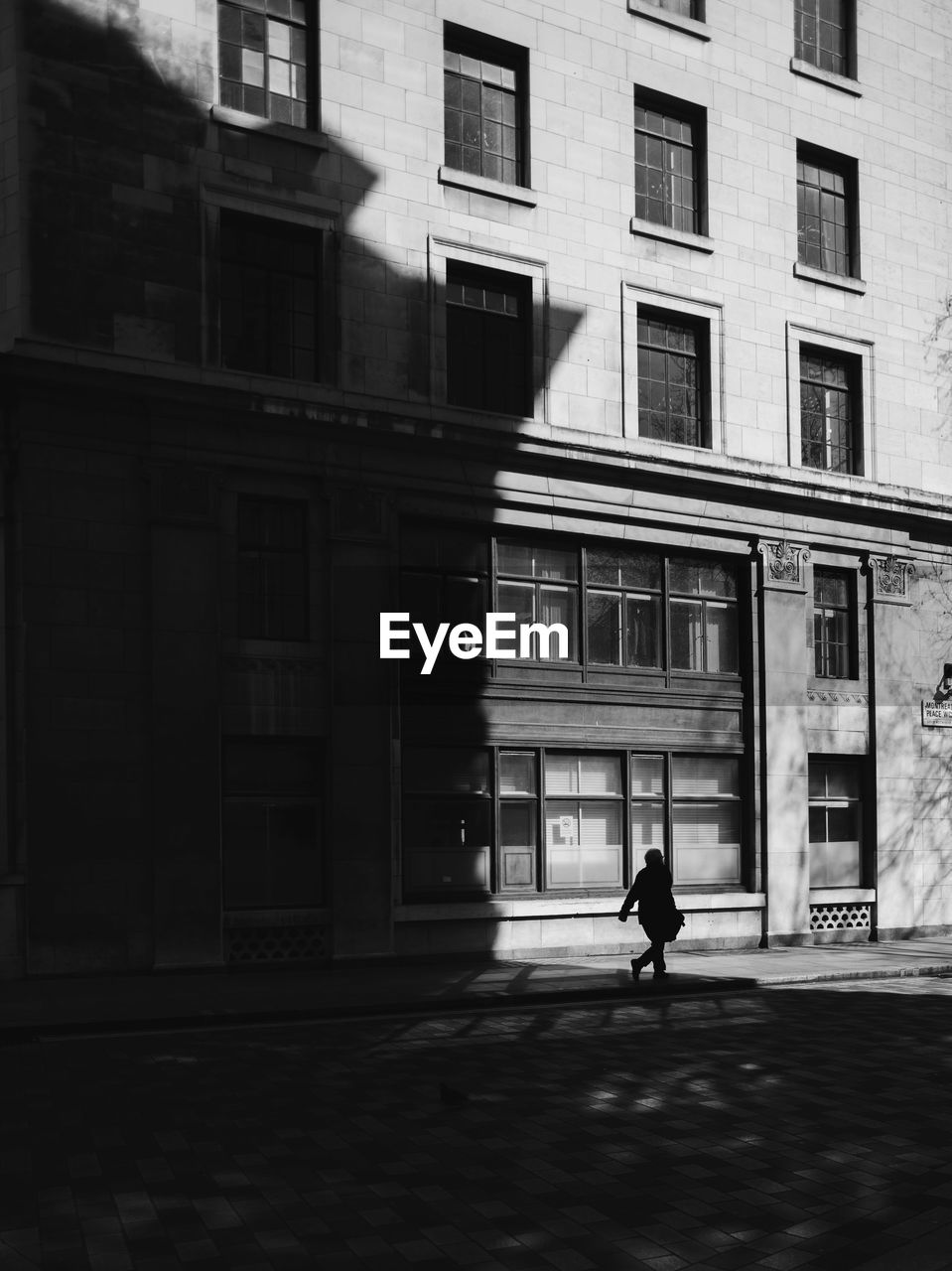 Silhouette man walking on footpath by building during sunny day