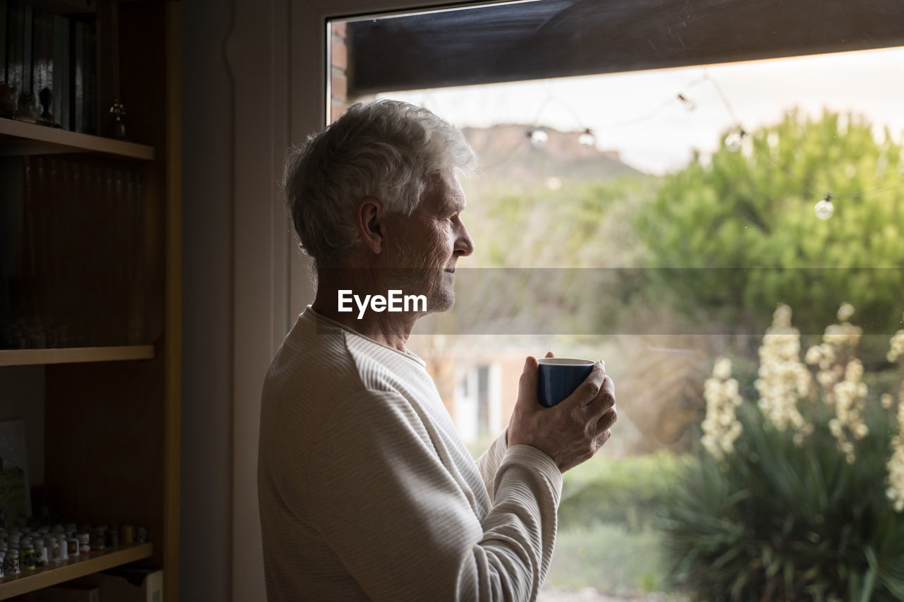 Senior man with coffee cup looking through window while standing at home