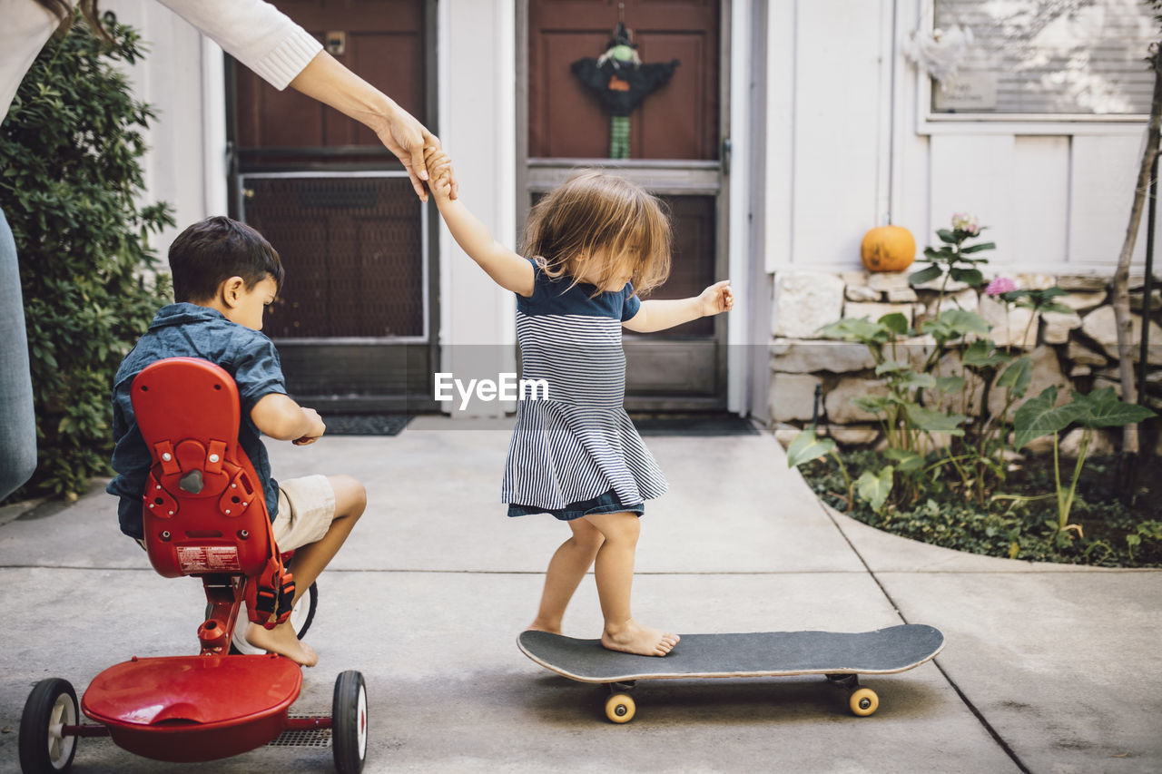 Daughter skateboarding with help of mother while son sitting on tricycle