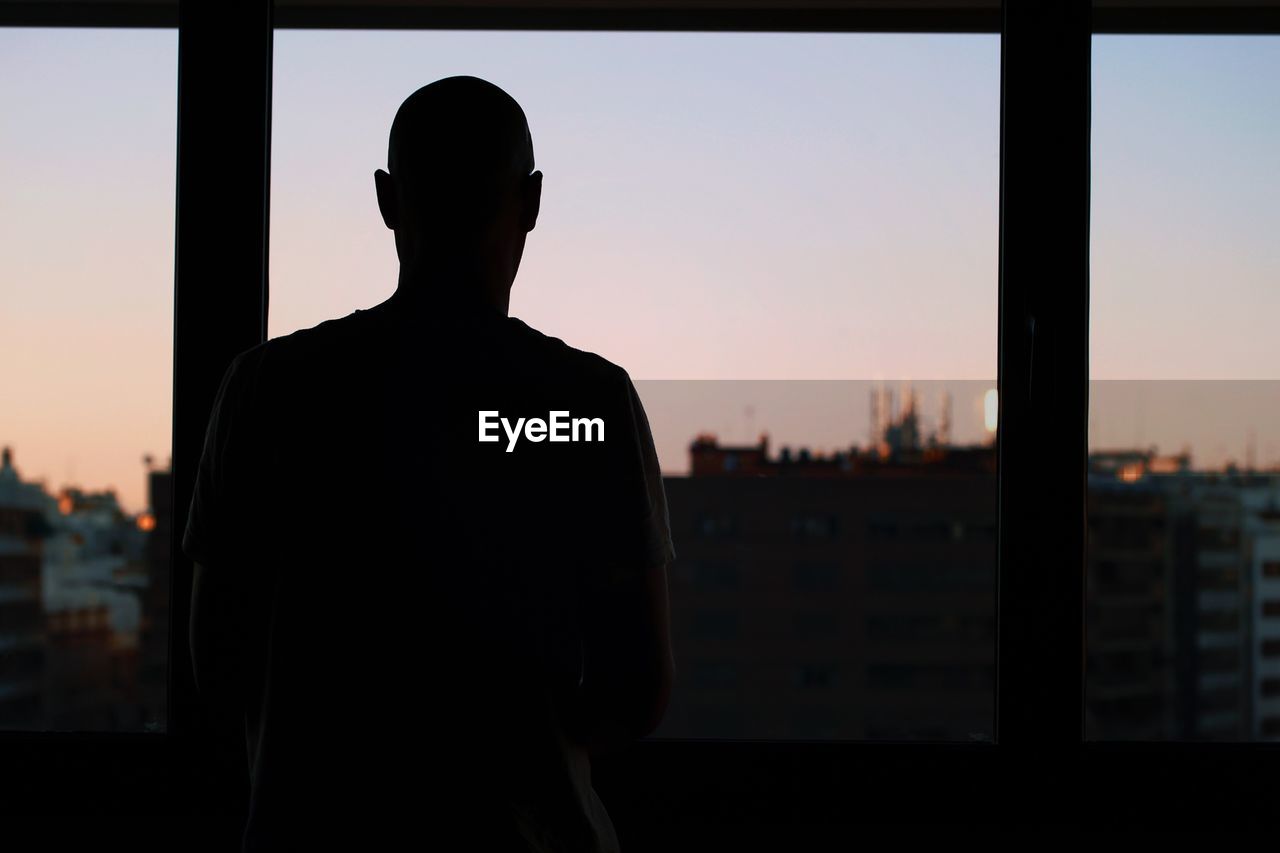 Rear view of silhouette man looking through window during sunset